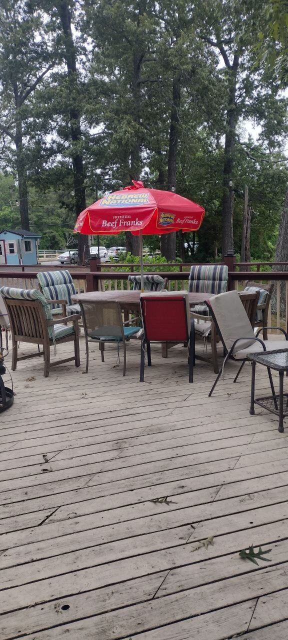 Pemberton Coffeehouse deck table under a red Hebrew National umbrella, weathered grey deck planks, oaks in the background
