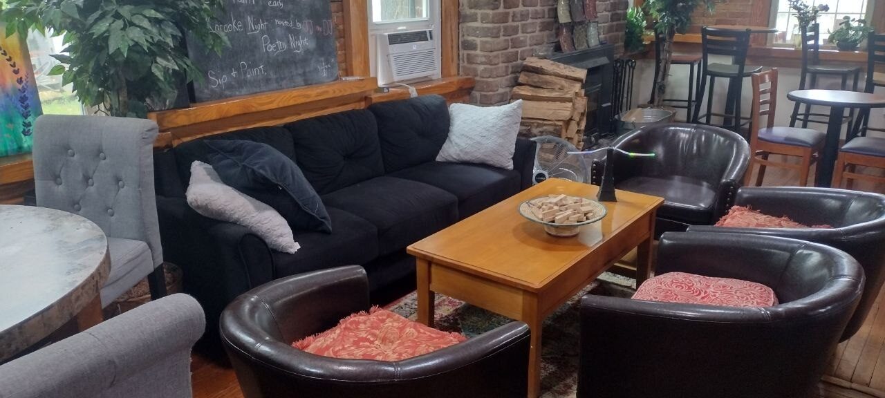 Pemberton Coffeehouse lounge corner — charcoal sofa, leather barrel chairs, events chalkboard listing Karaoke Night and Poetry Night