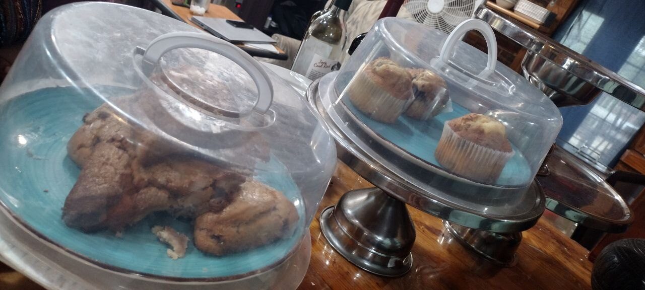 Pemberton Coffeehouse pastry case — glass-dome displays of chocolate-chip cookies and blueberry muffins on the wood bar