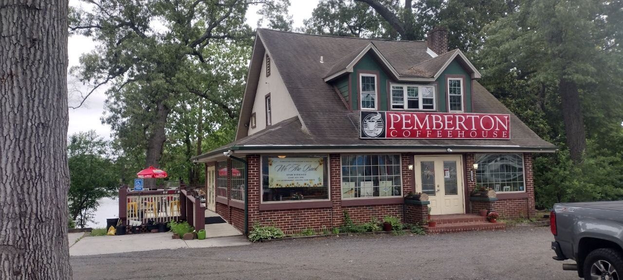 Pemberton Coffeehouse exterior — brick and green-trim building beside the pond with a 'We Are Back' sunflower banner in the window
