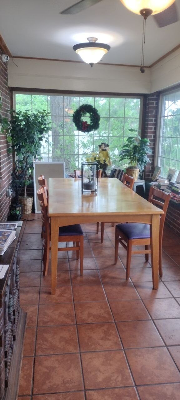 Pemberton Coffeehouse private dining sunroom — long blond-wood farmhouse table, wood-and-vinyl chairs, brick walls, windows wrapping the back